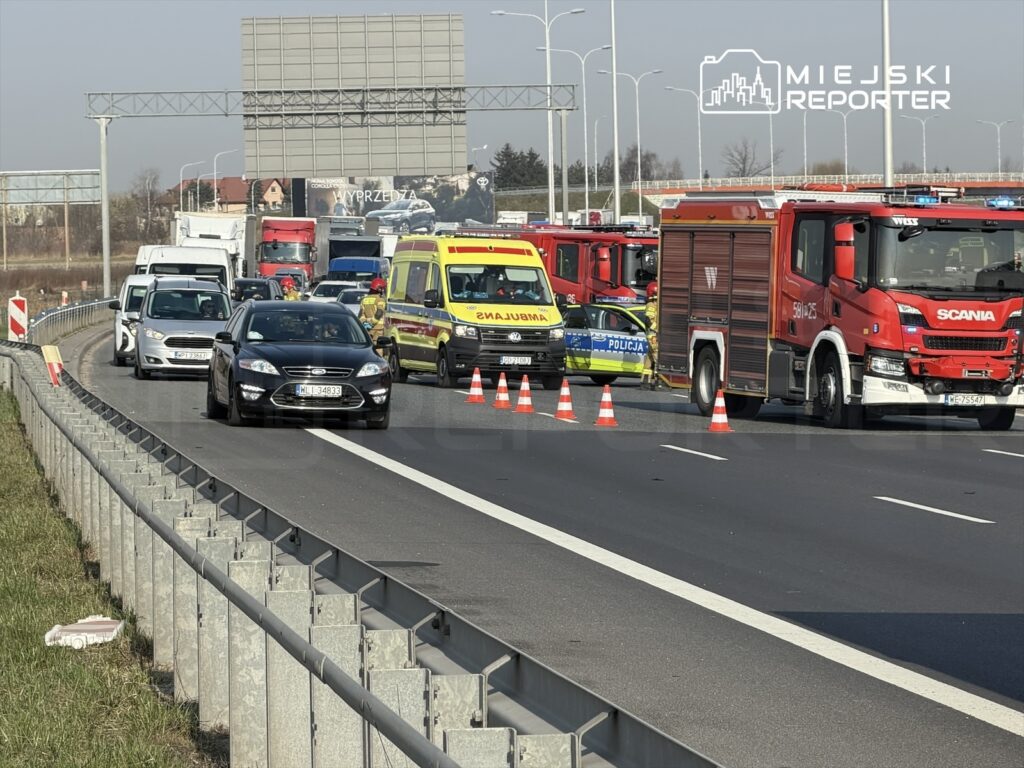 Na drodze stoją pojazdy ratunkowe, w tym karetka i wóz strażacki, a ruch jest spowolniony przez ustawione pachołki.