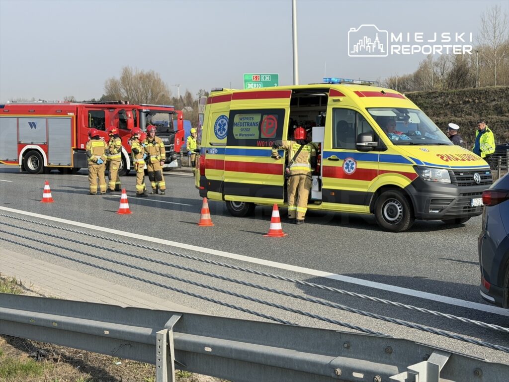 Zespół ratowników medycznych i strażaków pracuje przy wypadku na drodze, obok karetki i wozu strażackiego.