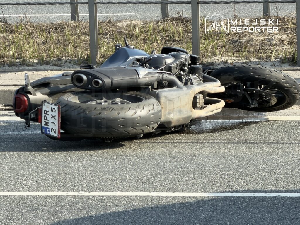 Motocykl leżący na boku na drodze, z widocznymi dużymi oponami i uszkodzeniami silnika.
