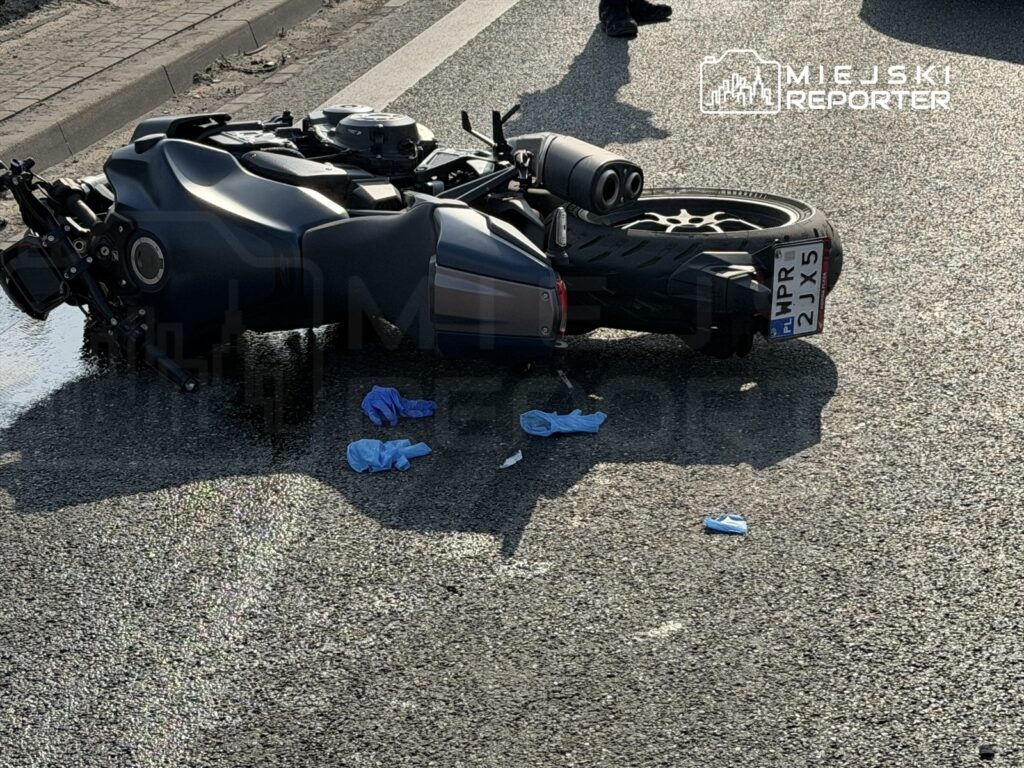 Motocykl leżący na boku na drodze, obok niebieskie rękawiczki i plama płynu.
