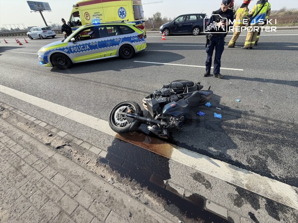 Motocykl leżący na drodze, obok policjanci i karetka, w tle samochody oraz oznaczenia drogi.
