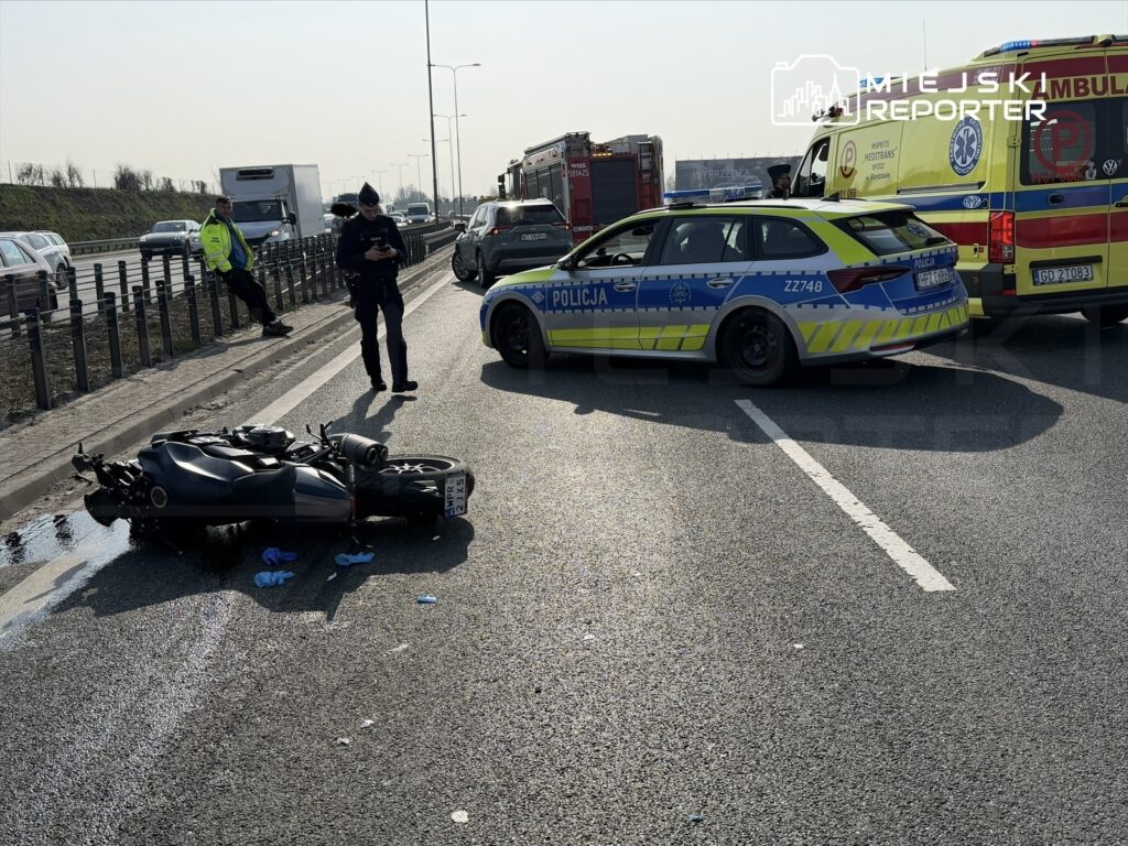 Motocykl leży na drodze, a obok stoją policjanci i karetka, w tle widać pojazdy w ruchu.