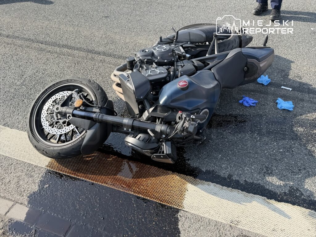 Motocykl leżący na drodze, wokół plama oleju oraz niebieskie rękawiczki ochronne.