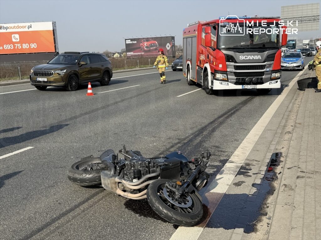 Motocykl leży na jezdni, obok widać czerwony wóz strażacki oraz samochód osobowy, a w tle billboard reklamowy.
