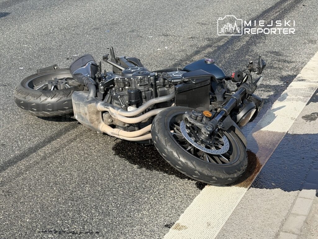 Motocykl leżący na boku na jezdni, z wyciekającym płynem, w otoczeniu asfaltu.