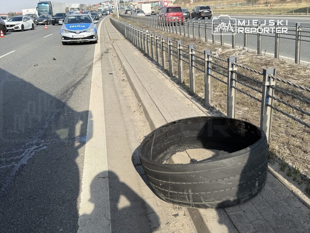 Na poboczu drogi leży uszkodzona opona, obok niej znajduje się radiowóz policyjny, a w tle widać zator drogowy.