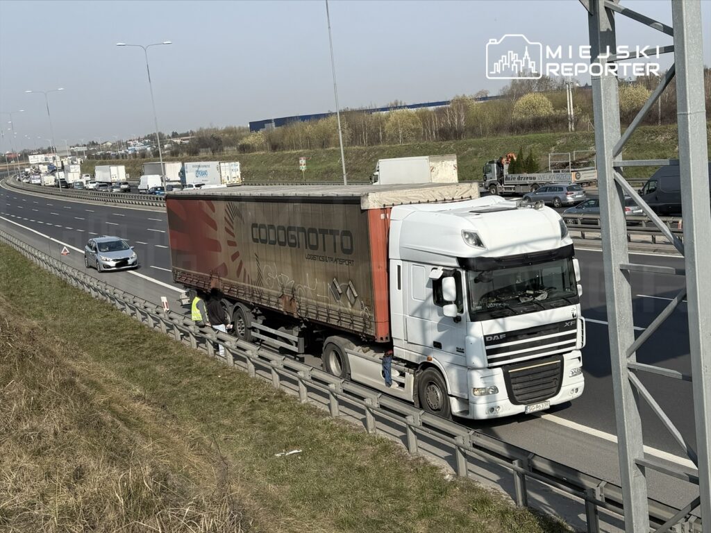 Czterech pracowników w kamizelkach odblaskowych sprawdza ciężarówkę na poboczu ruchliwej drogi ekspresowej.