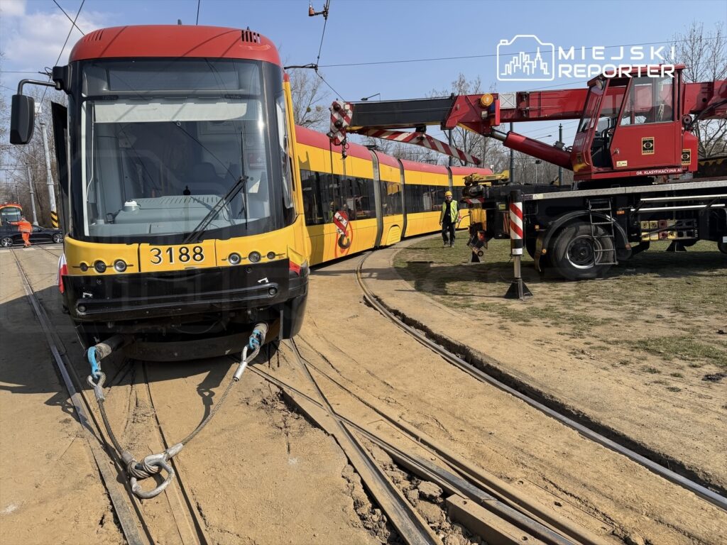 Tramwaj z numerem 3188 unieruchomiony na torach, holowany przez dźwig na placu budowy w słoneczny dzień.