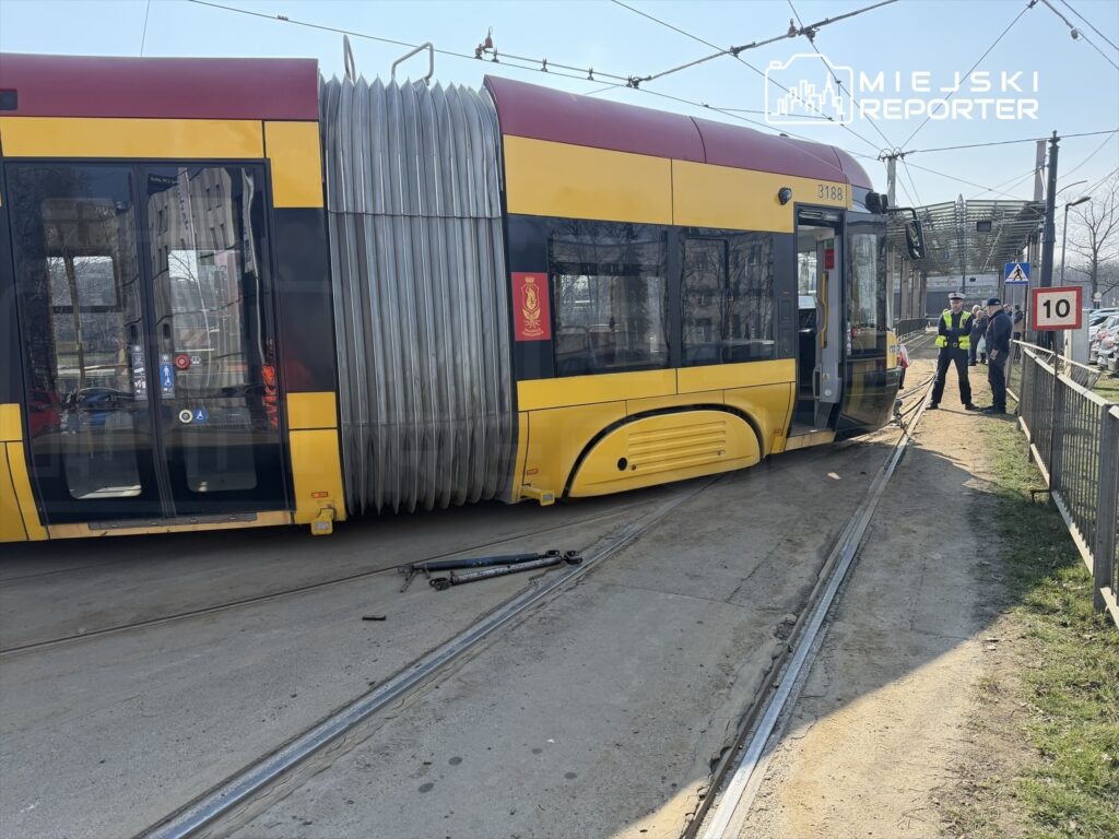 Tramwaj w żółto-czarnym malowaniu leży na boku obok torów, a obok stoją policjanci rozmawiający z mężczyzną.