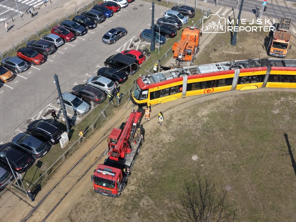 Tramwaj z uszkodzeniami stoi na torach, obok pracują robotnicy i wóz strażacki, w tle zaparkowane samochody.