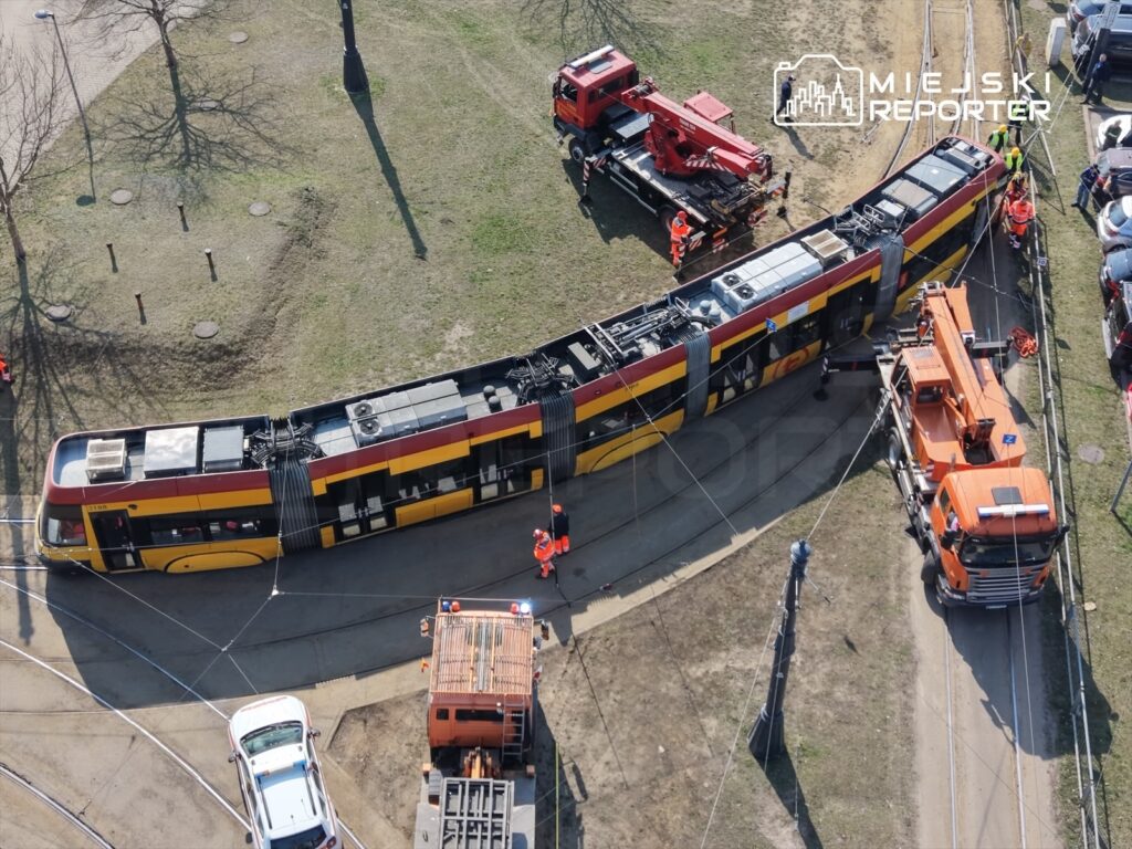 Tramwaj w kolorze żółto-czerwonym stoi na torach, wokół pracują robotnicy w pomarańczowych ubraniach.