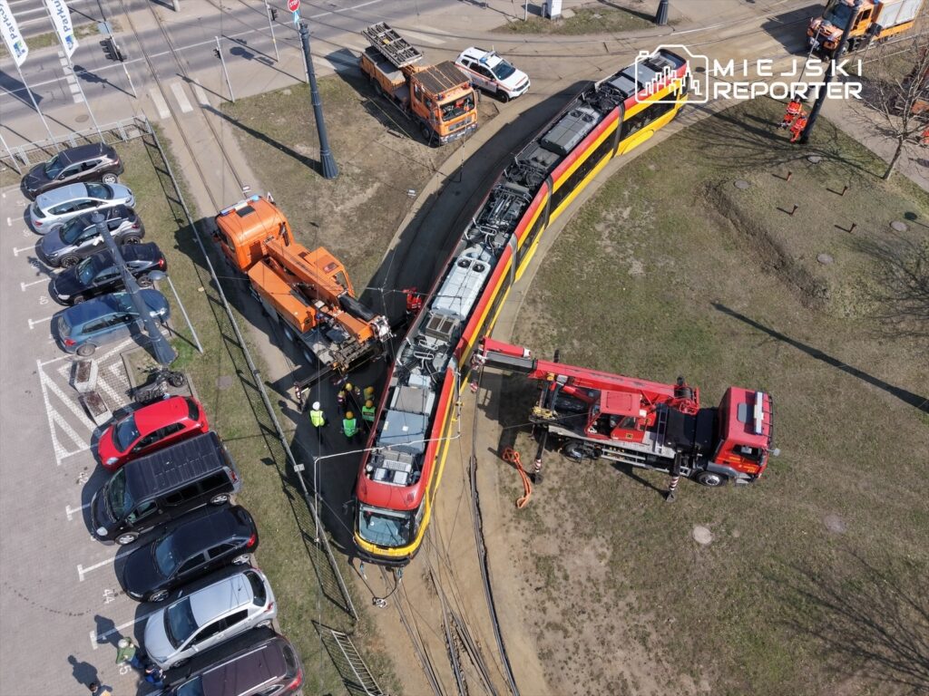 Ekipa techniczna naprawia tramwaj za pomocą ciężkiego sprzętu na torowisku w miejskim otoczeniu, obok zaparkowanych samochodów.