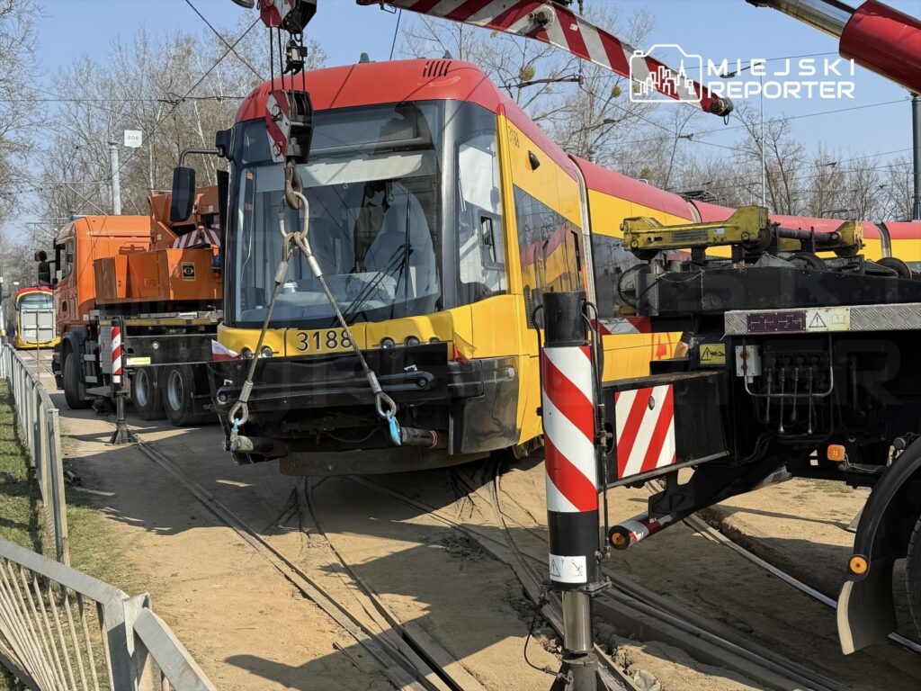 Tramwaj z numerem 3188 unosi się nad torami, obok ciężarówki z podnośnikiem w otoczeniu budowy.