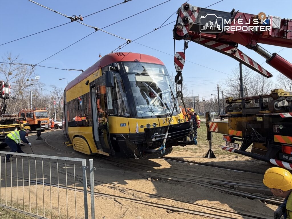 Tramwaj w żółto-czerwonym kolorze jest podnoszony przez dźwig na torowisku, a pracownicy w kamizelkach odblaskowych go obsługują.