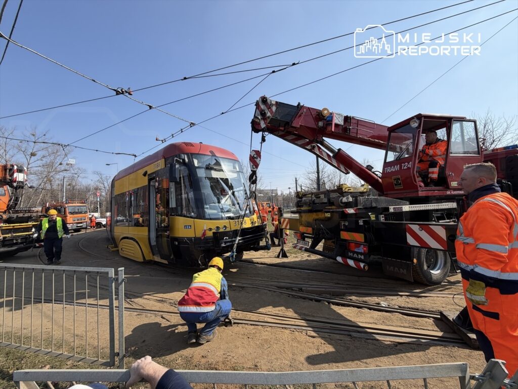 Pracownicy w kamizelkach odblaskowych używają dźwigu do podnoszenia tramwaju na torach.