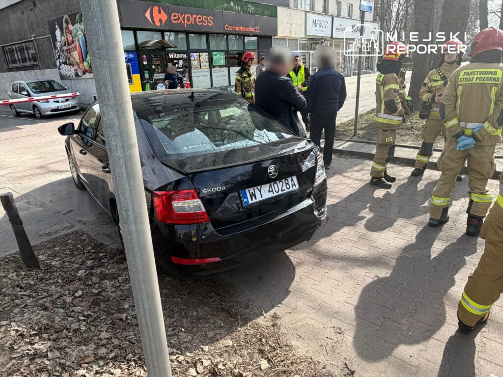 Czterech strażaków w mundurach rozmawia z mężczyzną obok uszkodzonego czarnego samochodu na chodniku przy sklepie.