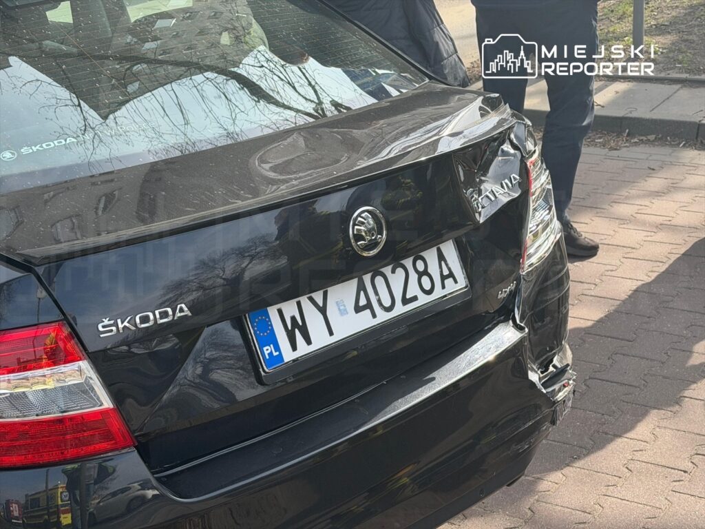 Czarny samochód marki Škoda z uszkodzoną tylną częścią stoi na chodniku, obok grupy ludzi.