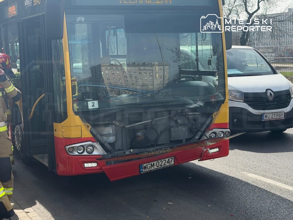 Autobus miejski z uszkodzoną przednią częścią stoi na ulicy, obok pracują strażacy w mundurach.