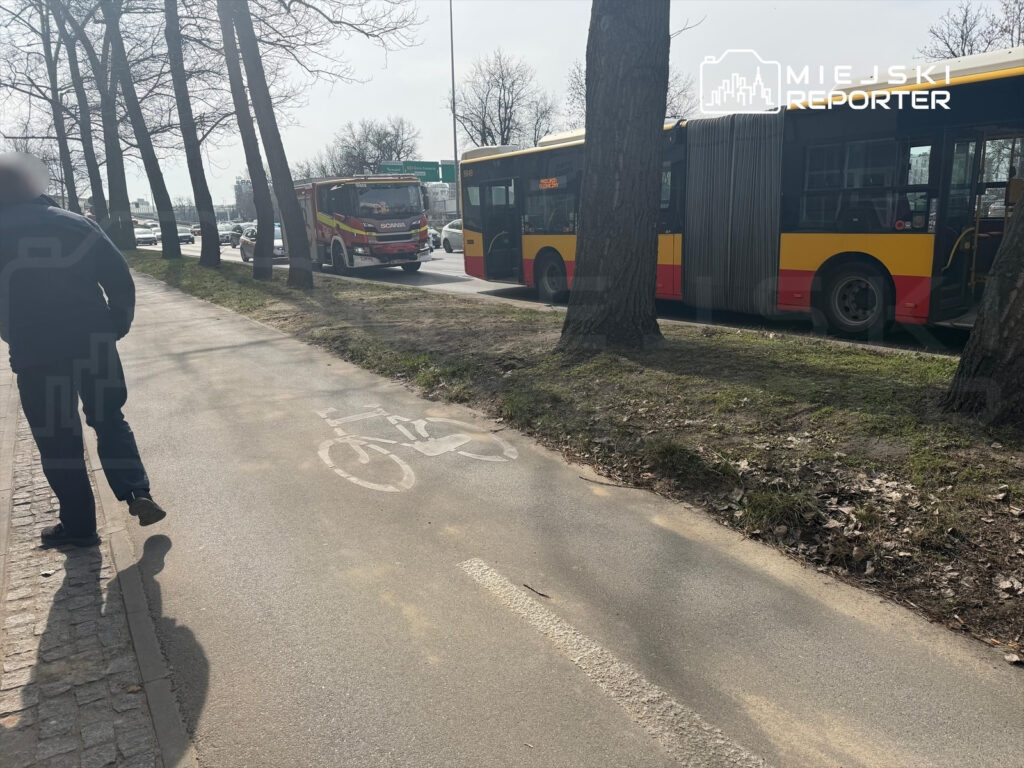 Mężczyzna spacerujący chodnikiem obok ruchliwej drogi, obok przystanku z autobusami i widocznymi oznaczeniami dla rowerzystów.