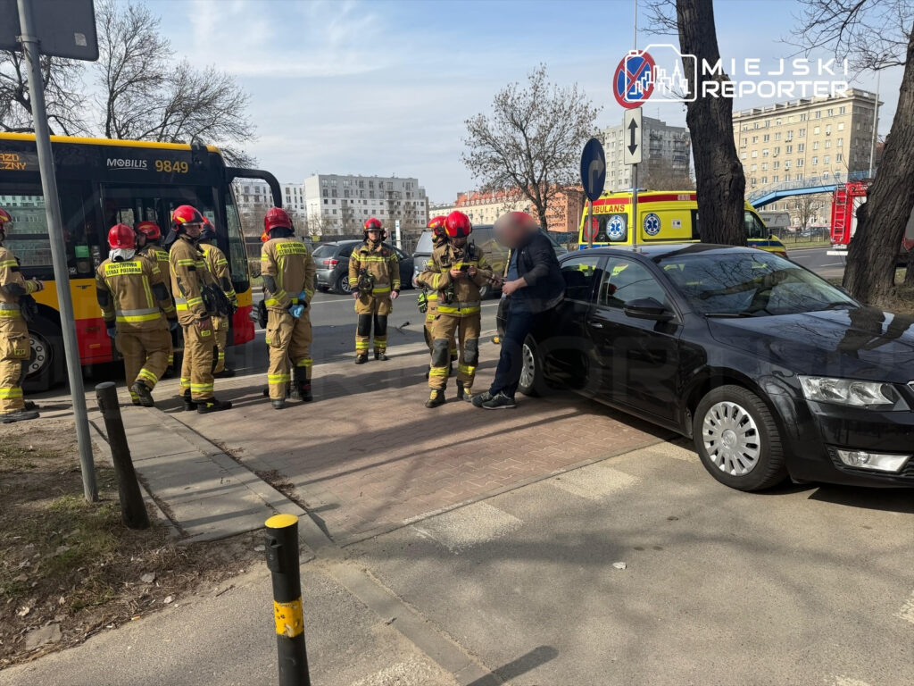 Grupa strażaków w mundurach omawia sytuację obok czarnego samochodu osobowego na chodniku.