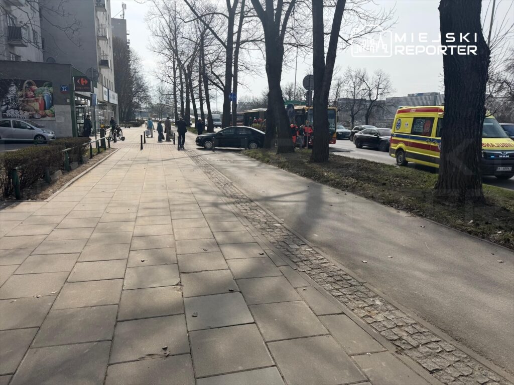 Grupa ludzi przechodzi chodnikiem obok nowoczesnego biurowca, a w tle widoczne są pojazdy ratunkowe.