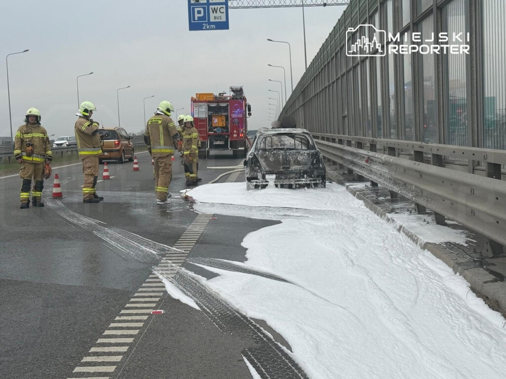 Czterech strażaków w mundurach na drodze obok samochodu objętego pożarem, pokrytego pianą gaśniczą.
