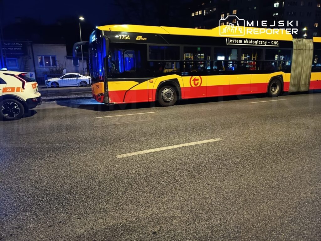 Autobus miejski w barwach żółto-czerwonych zatrzymany na ulicy, obok stoi radiowóz policyjny z włączonymi sygnałami świetlnymi.