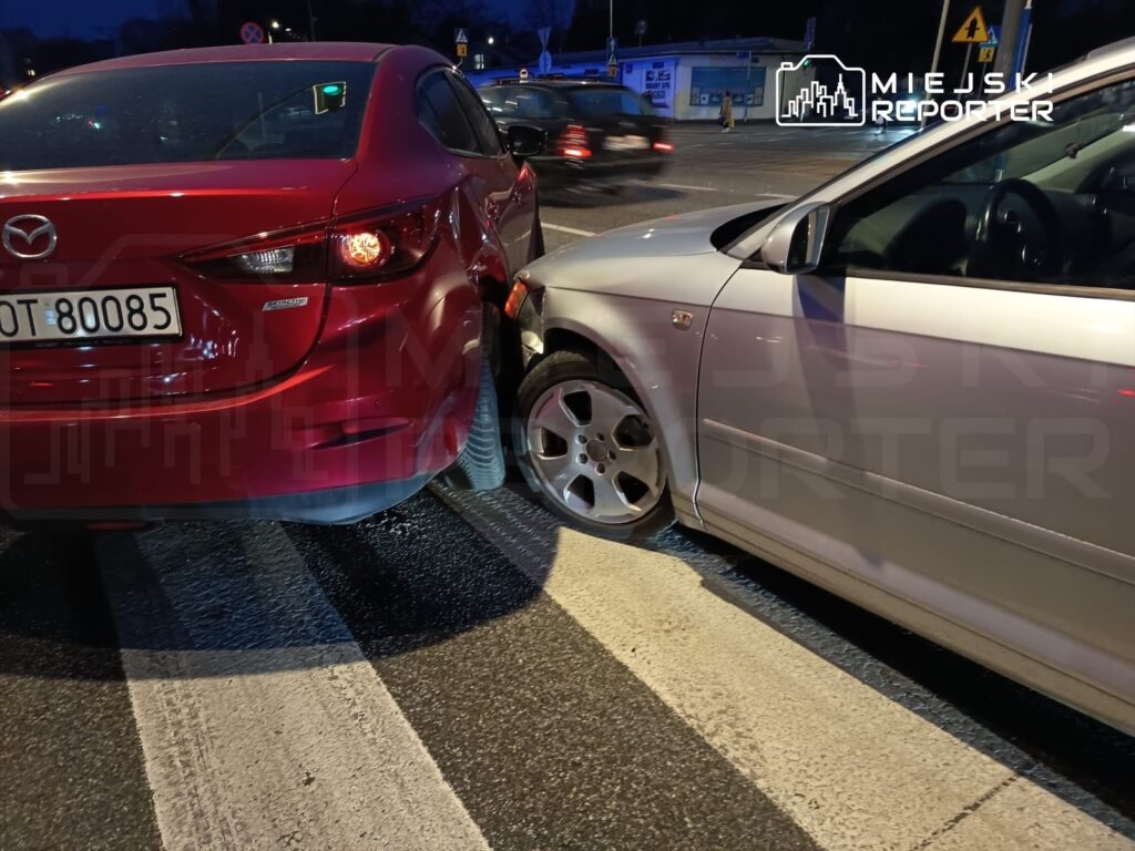 Czerwony samochód osobowy z uszkodzeniami tyłu zderza się z szarym pojazdem na przejściu dla pieszych w nocy.