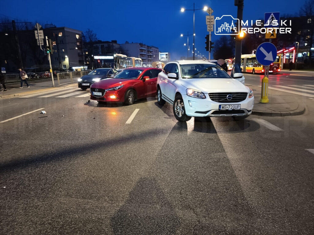 Dwa uszkodzone samochody osobowe na skrzyżowaniu w mieście, w tle tramwaj i piesi na chodniku.