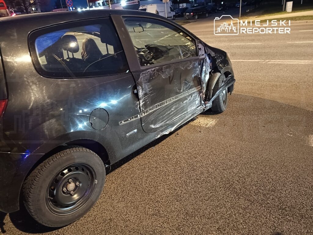 Czarny samochód osobowy z uszkodzeniami na bocznej części stoi na drodze, obok zaparkowanych pojazdów.