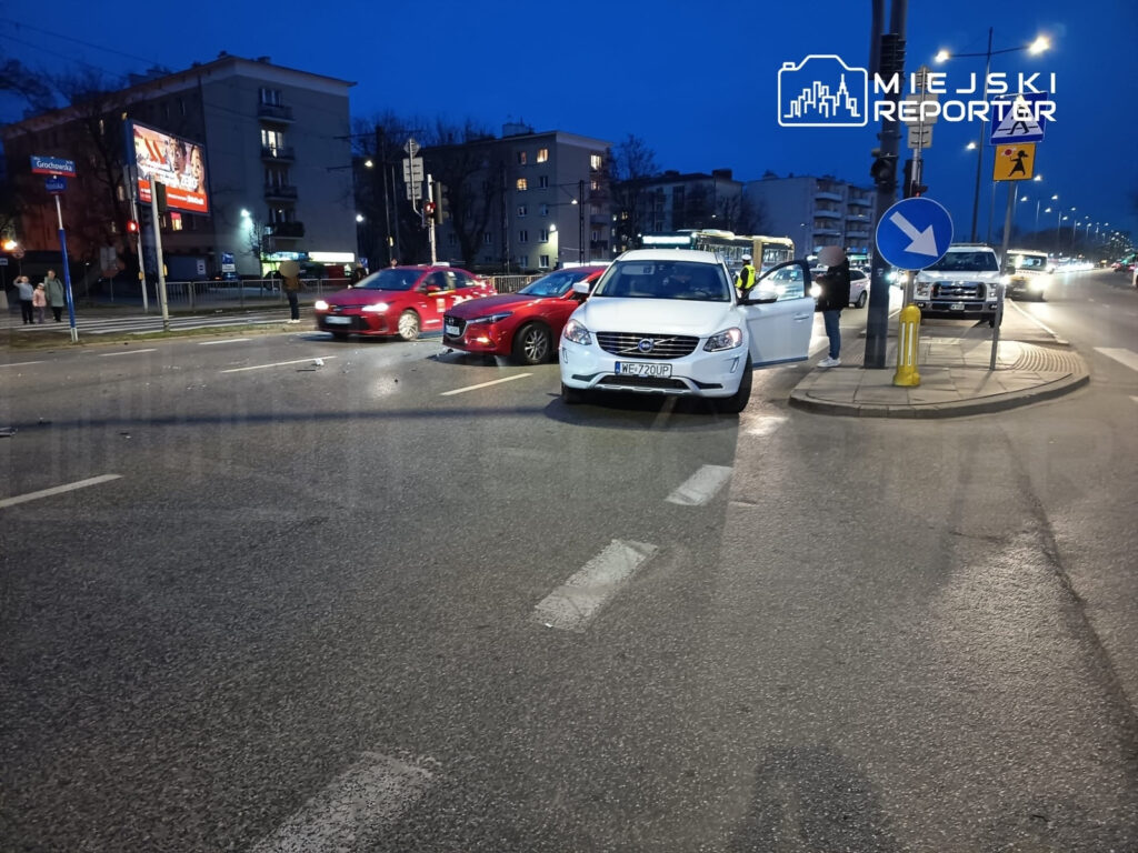 W nocy na skrzyżowaniu ulic stoją dwa uszkodzone samochody: czerwony i biały, obok nich widać ludzi oraz sygnalizację świetlną.