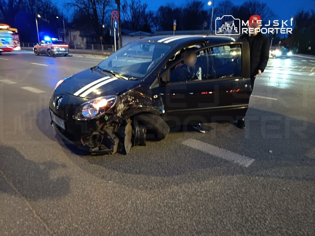 Czarny samochód osobowy z uszkodzeniem przedniego koła stoi na skrzyżowaniu, obok widać radiowóz i karetkę.
