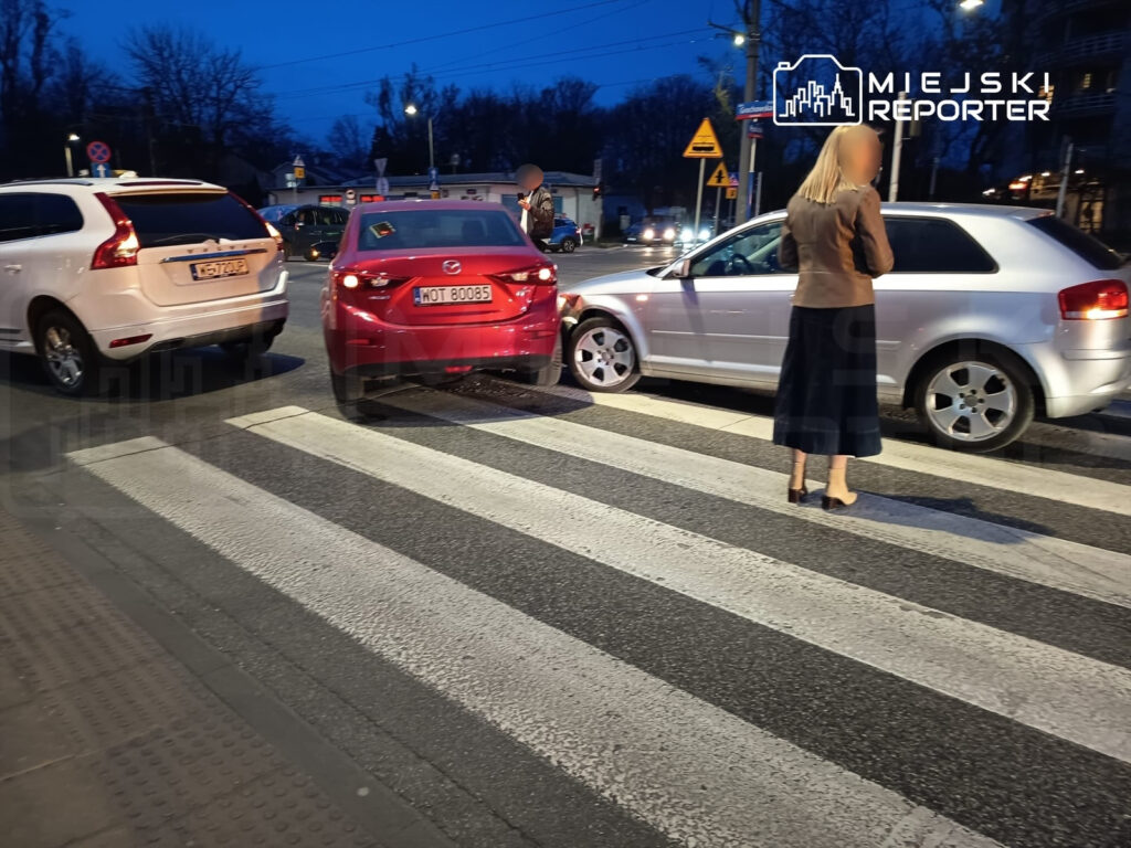Kobieta w brązowej kurtce stoi na przejściu dla pieszych obok zaparkowanych samochodów w miejskim otoczeniu.