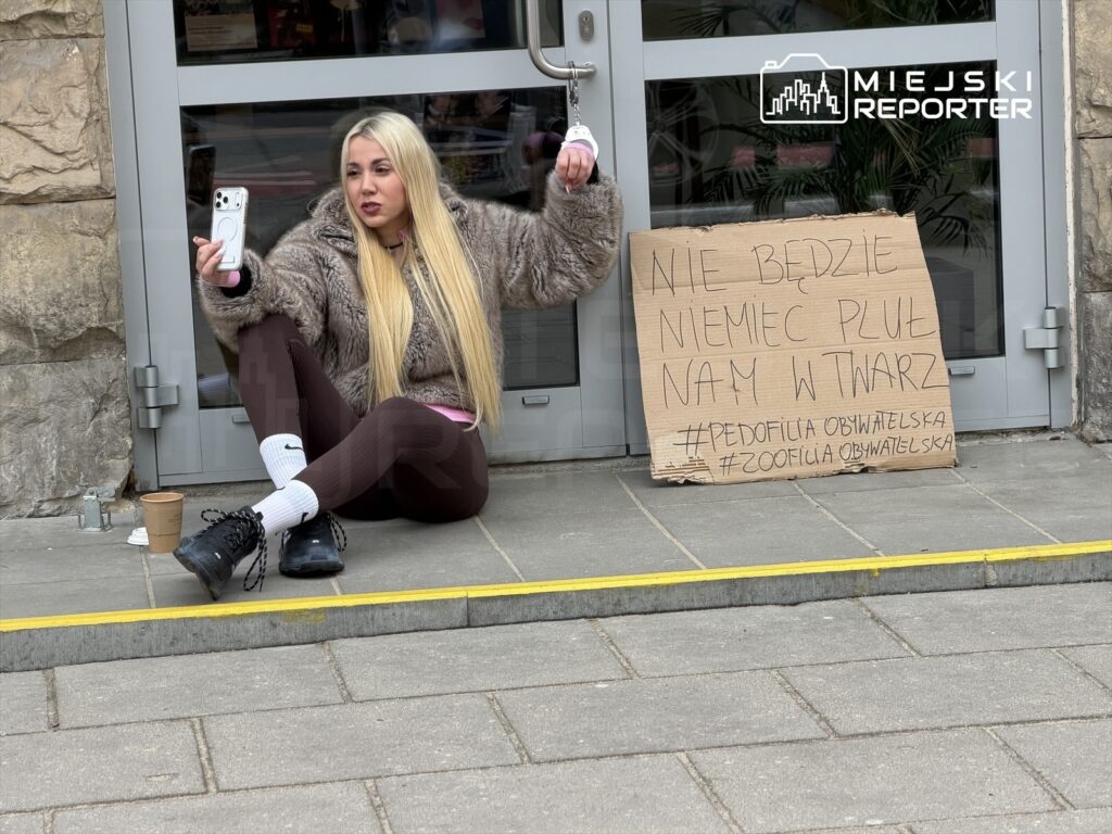 Kobieta w futrze siedzi na chodniku, trzyma telefon i robi zdjęcie, obok niej karton z napisem: "NIE BĘDZIE NIEMIEC PLUŁ NAM W TWARZ".