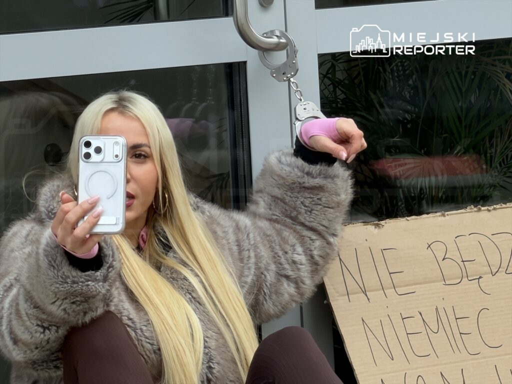 Kobieta w futrzanej kurtce trzyma telefon i robi selfie, siedząc na ziemi obok kartonu z napisem „NIE BĘDZIE NIEMIEC W MONACHIUM”.