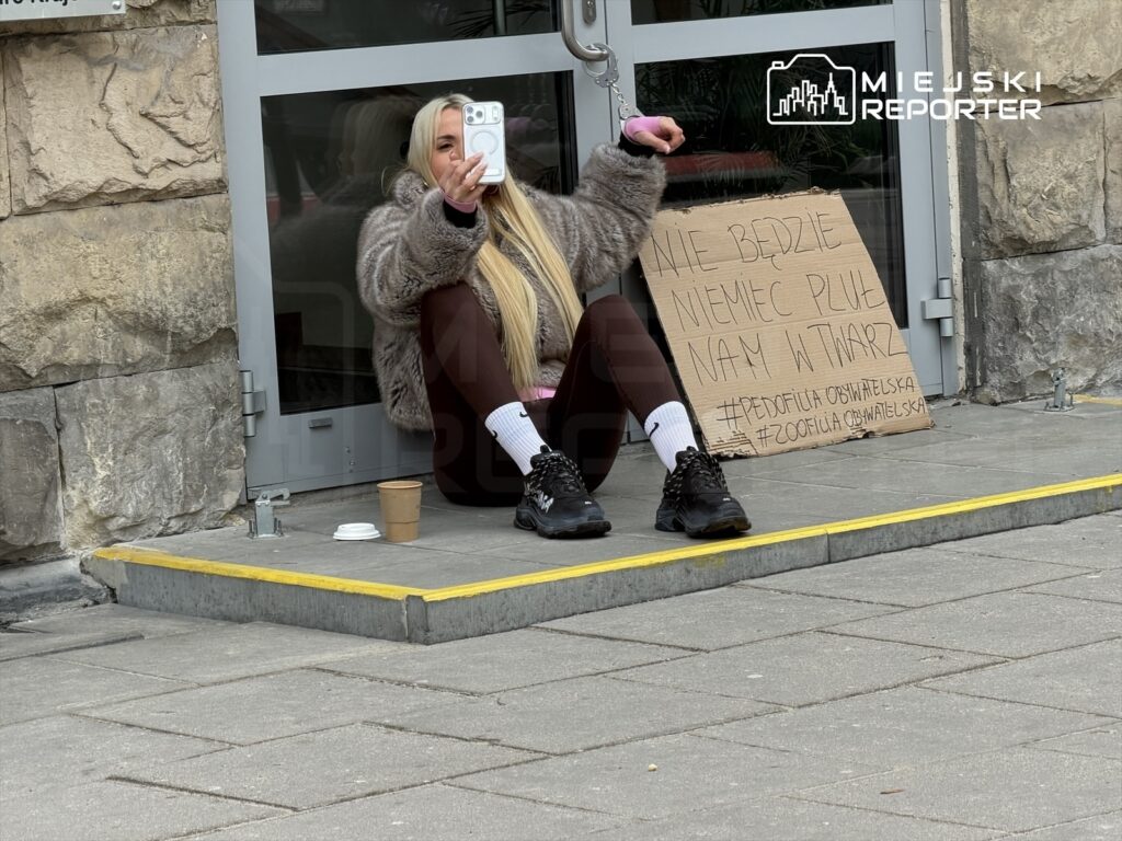 Kobieta w futrzanej kurtce siedzi na schodach, trzymając telefon i pozując do zdjęcia, obok niej leży karton z napisem: "Nie będzie Niemiec pluje nam w twarz".