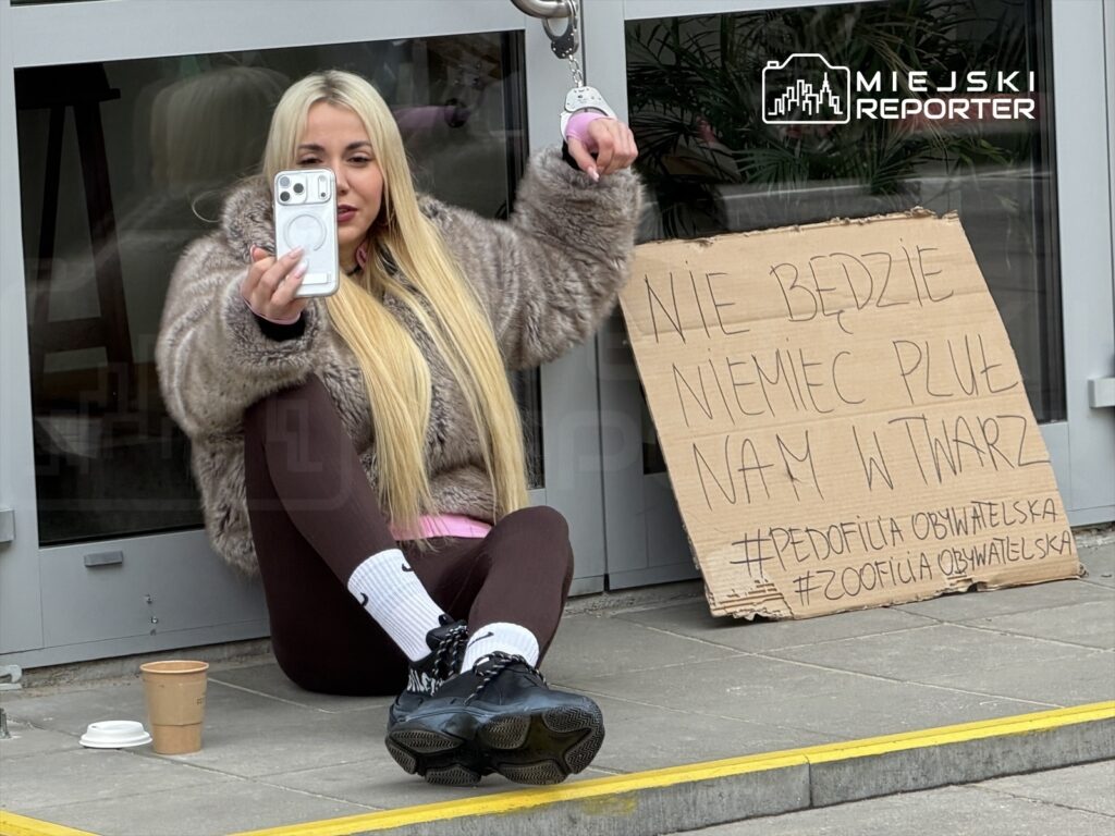 Kobieta w futrzanej kurtce siedzi na schodach, trzymając telefon i robiąc selfie, obok niej transparent z napisem: "NIE BĘDZIE NIEMIEC PLUŁ NAM W TWARZ".