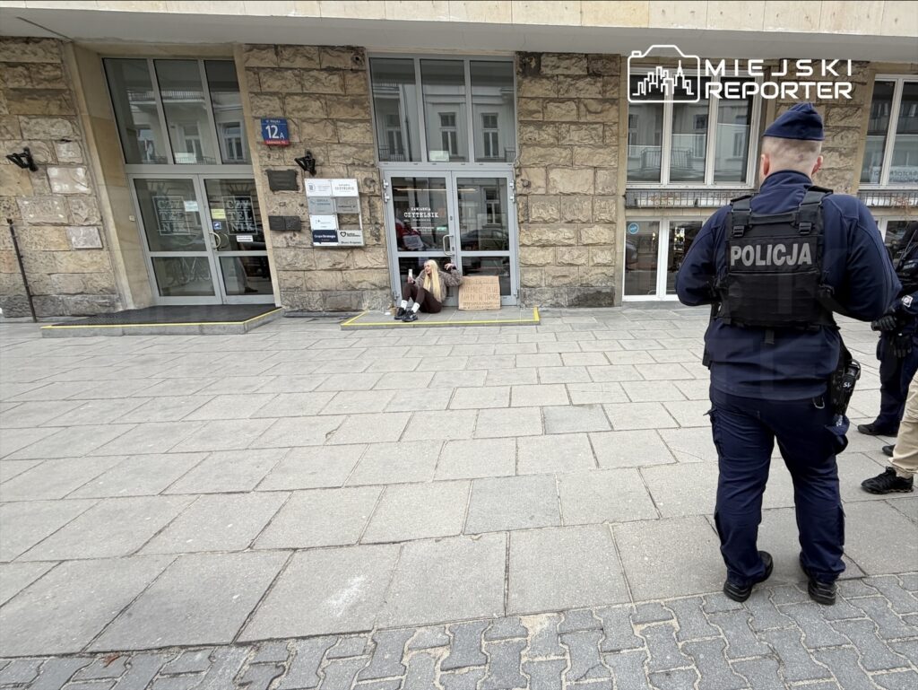 Mężczyzna siedzący na chodniku przed budynkiem z napisem, obok dwóch policjantów rozmawiających ze sobą.