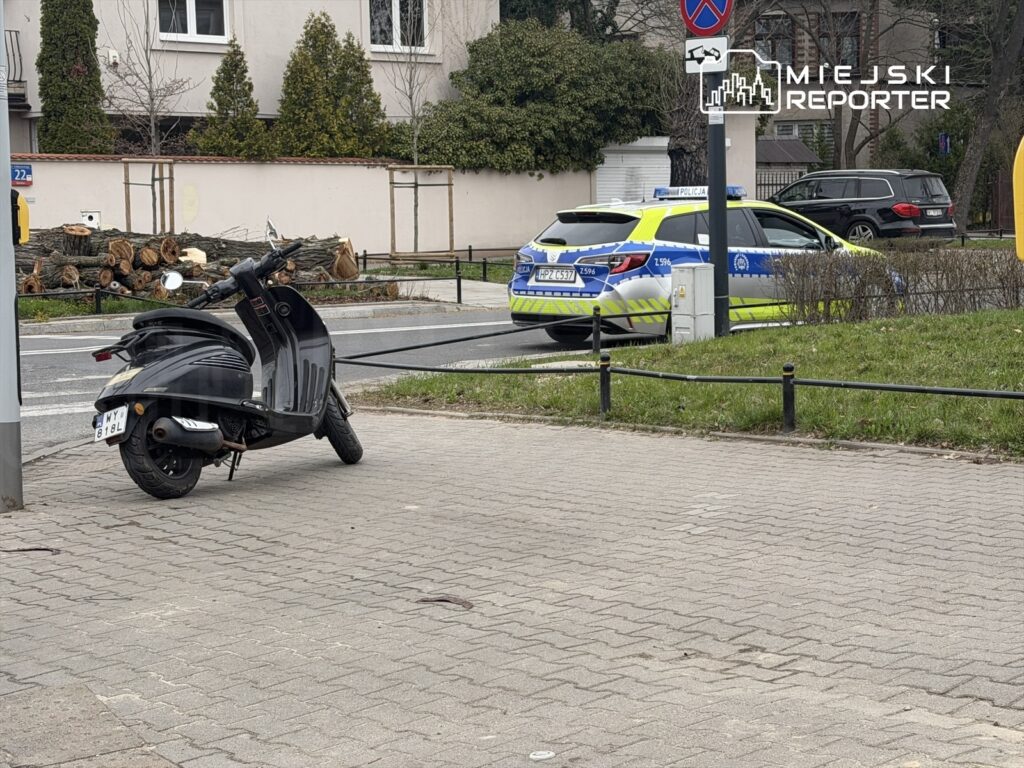 Czarny skuter zaparkowany na chodniku obok policyjnego samochodu w nieoznakowanej strefie.