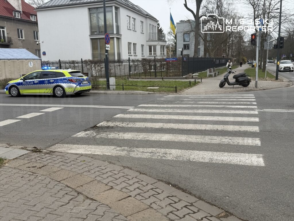 Policyjny radiowóz z włączonymi sygnałami świetlnymi stoi obok przejścia dla pieszych na miejskiej ulicy.