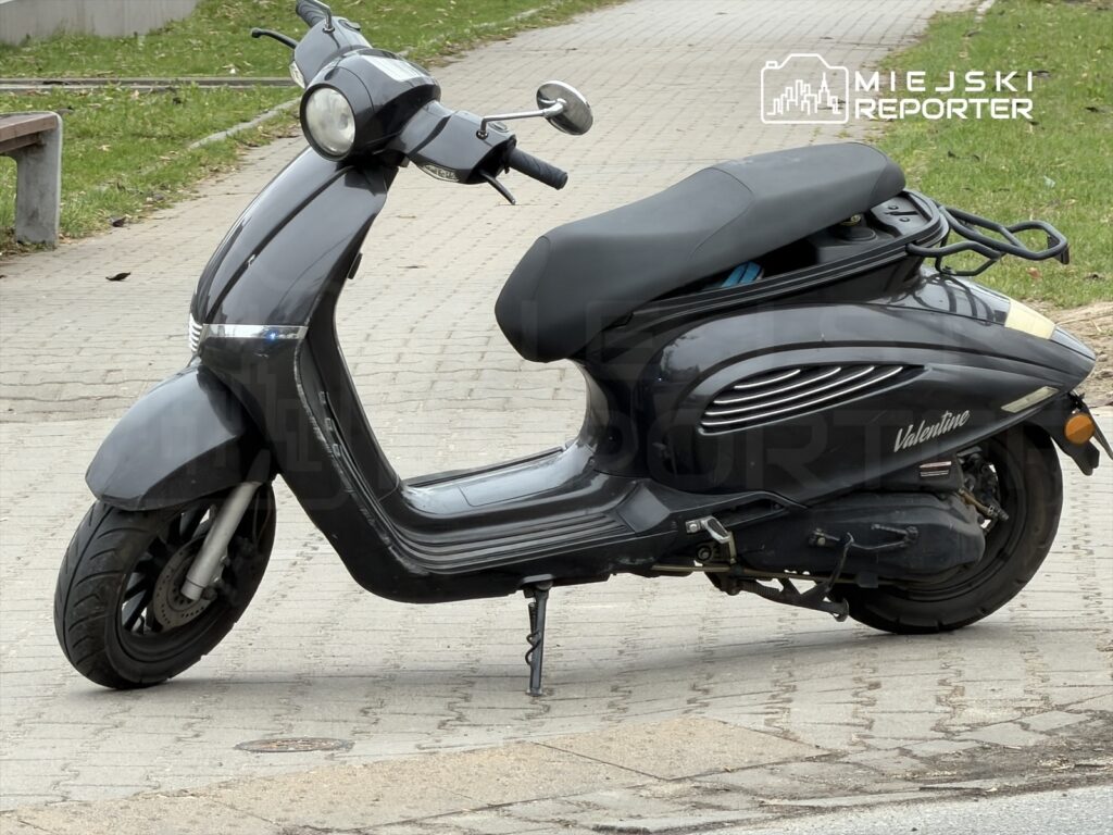 Czarny skuter zaparkowany na chodniku obok trawnika, z widocznymi detalami na nadwoziu.