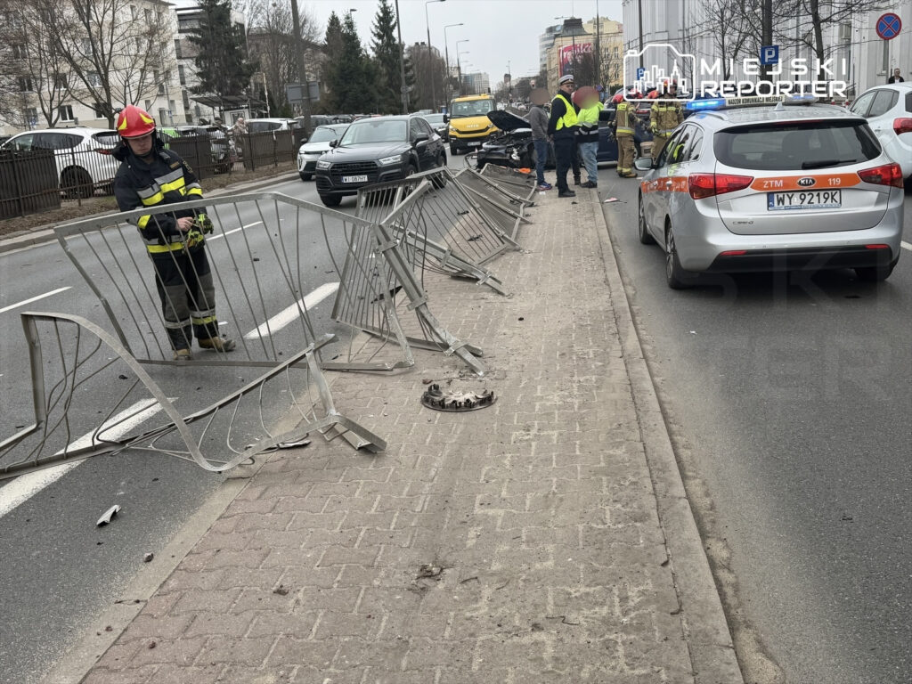 Strażak w mundurze bada uszkodzone barierki na jezdni, obok stoi uszkodzony pojazd oraz radiowóz policyjny.