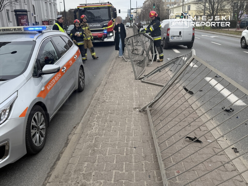 Na jezdni stoją strażacy i policjanci, obok uszkodzone bariery ochronne. W tle widać zaparkowany radiowóz.