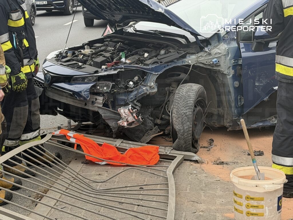 W pobliżu uszkodzonego samochodu stoją strażacy w uniformach, a na drodze leży pomarańczowy płaszcz ostrzegawczy.