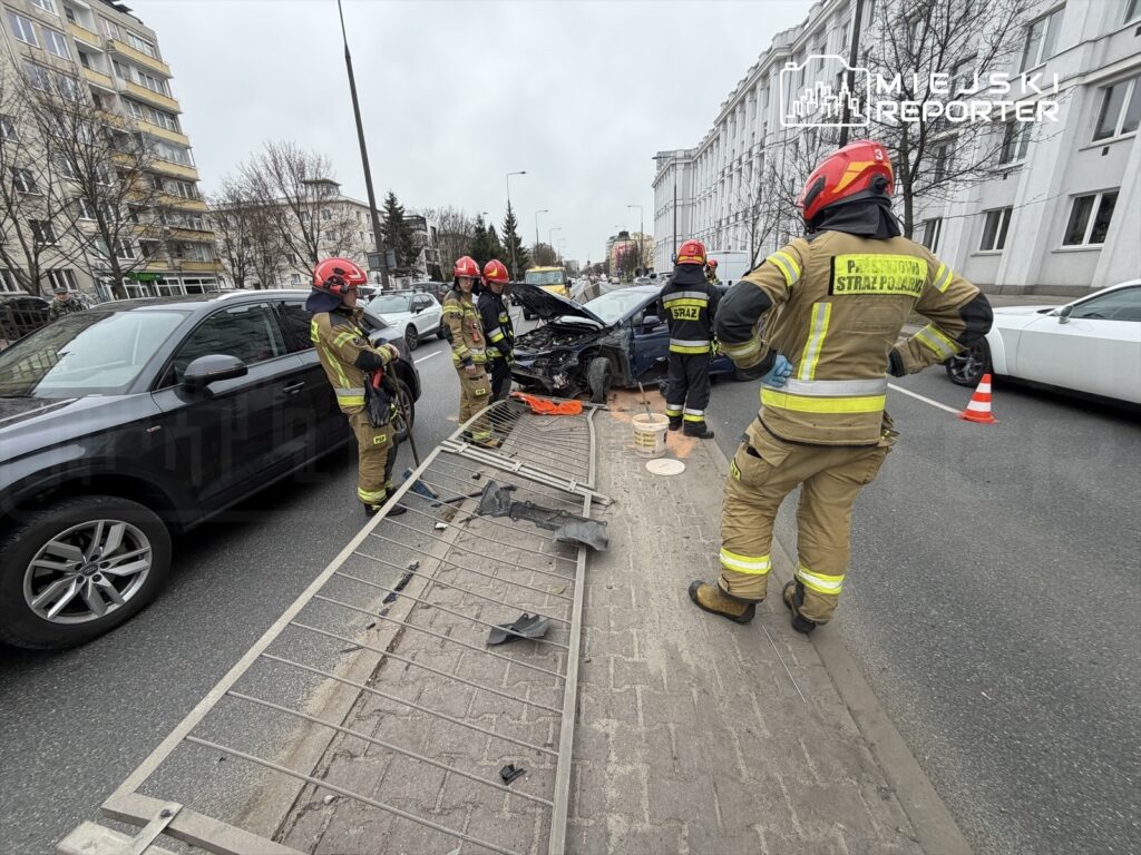 Czterech strażaków w hełmach ocenia uszkodzenia po wypadku drogowym na ruchliwej ulicy, obok rozbitego samochodu.