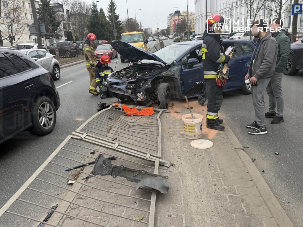 Ratownicy w kolorowych mundurach sprawdzają uszkodzony samochód na ruchliwej ulicy, wokół widać inne pojazdy.
