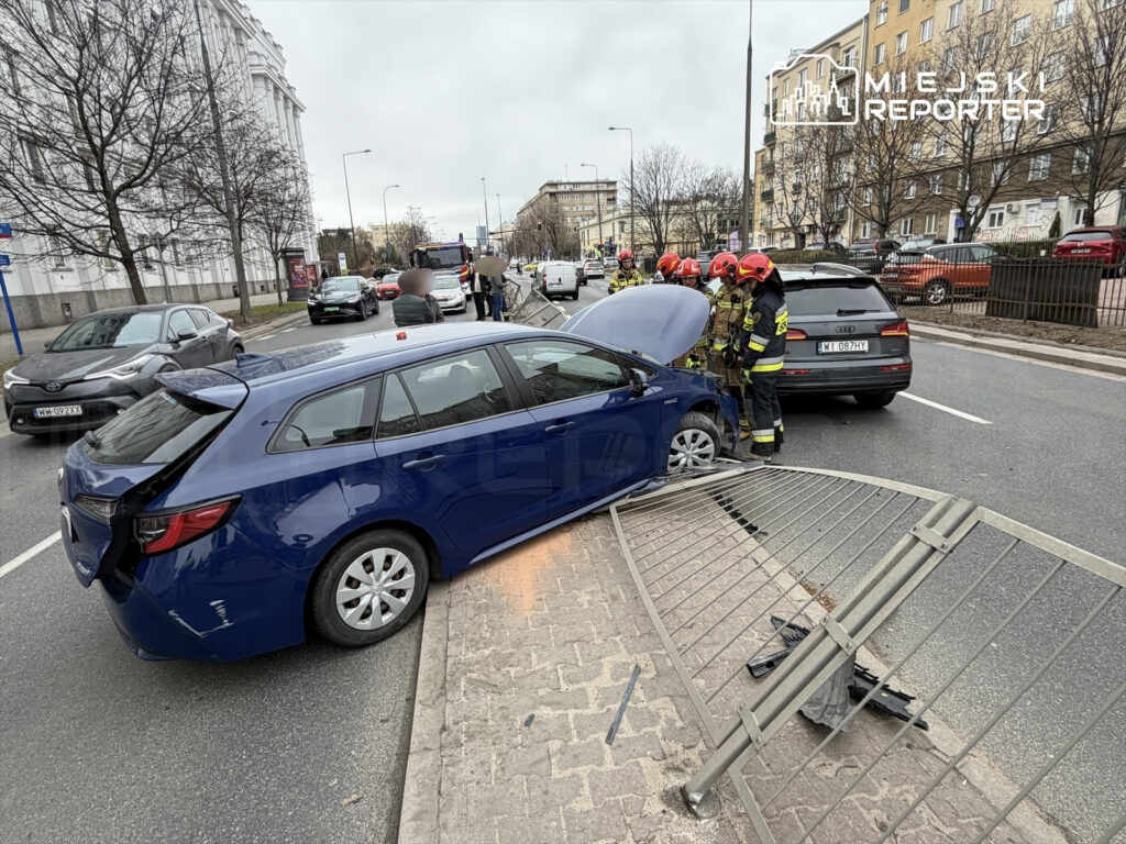 Niebieski samochód osobowy uderzył w ogrodzenie, a obok stoją strażacy w mundurach, interweniujący na miejscu zdarzenia.