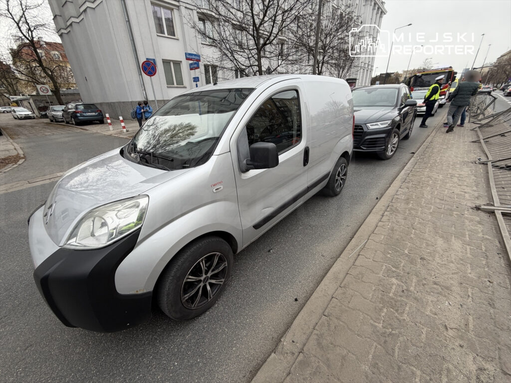 Srebrny samochód dostawczy zaparkowany na ulicy obok dwóch innych pojazdów, w tle widoczny znak zakazu parkowania.