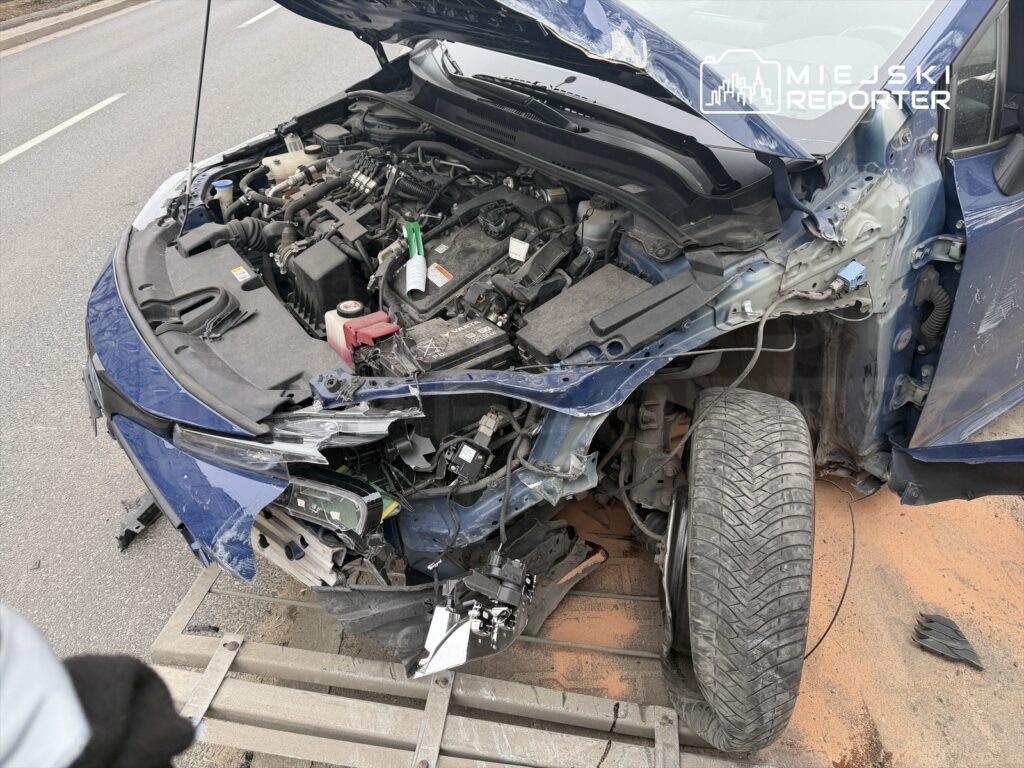 Uszkodzony samochód osobowy z otwartą maską, odsłaniającym silnik, stoi na poboczu drogi.