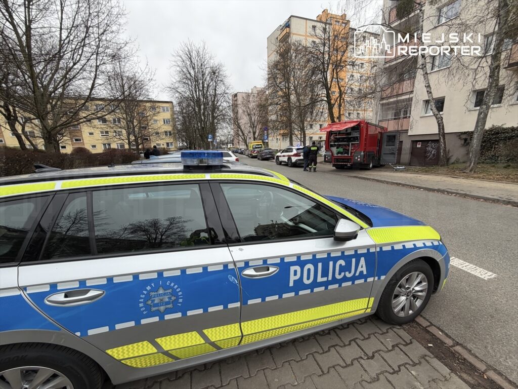 Radiowóz policyjny zaparkowany na ulicy, obok strażacki wóz i dwie osoby w mundurach, w tle bloki mieszkalne.